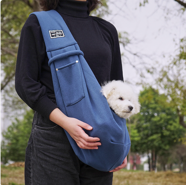 Person holding a blue pet sling with a small white dog peeking out, outdoors.