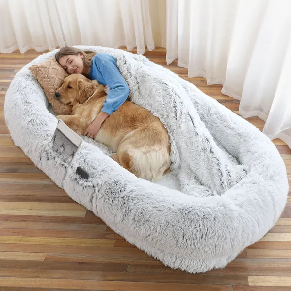 Woman hugging a large fluffy dog bed with a golden retriever inside on a wooden floor.