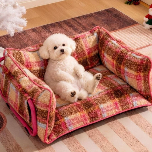Small white dog sitting on a checkered pet bed in a room with Christmas decorations.