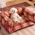 Small white dog sitting on a checkered pet bed in a room with Christmas decorations.