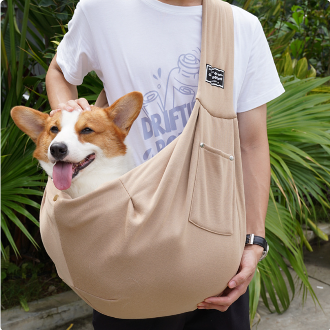 Dog in a beige sling held by a person with green foliage in the background
